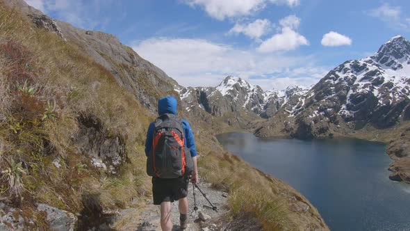 Follow, hiker walks above alpine lake, snow capped mountain landscape, Routeburn Track New Zealand