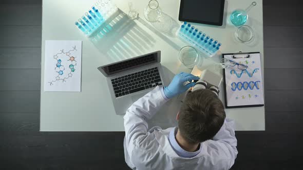 Scientist Viewing Samples Under Microscope and Writing Results on Laptop in Lab