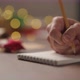 Slow Motion Orbit Shot Man Writes Christmas Gifts Notepad Pencil Under Warm Light Evening - VideoHive Item for Sale