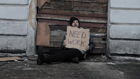 Homeless Man Sits and Holds Piece of Cardboard with Inscription Need Work