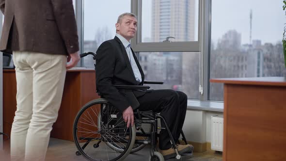 Side View of Sad Disabled Senior Man in Wheelchair Looking Out the Window in Office and Talking to