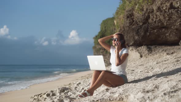 Woman Freelancer in Sunglasses Works on Beach with Laptop and Talks on Phone