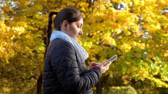 Young Woman in a Black Jacket and a Gray Scarf is Typing a Message on the Phone Against the