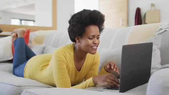 Happy african american woman laying on sofa using laptop