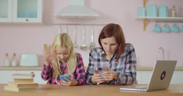 Blond Schoolgirl and Young Mother Browsing Internet By Mobile Phones Sitting at Home at Pink Kitchen