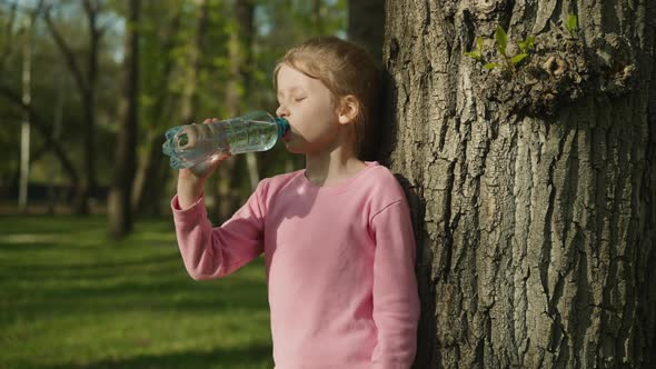 Tired Little Girl Drinks Water Leaning on Old Tree in Park