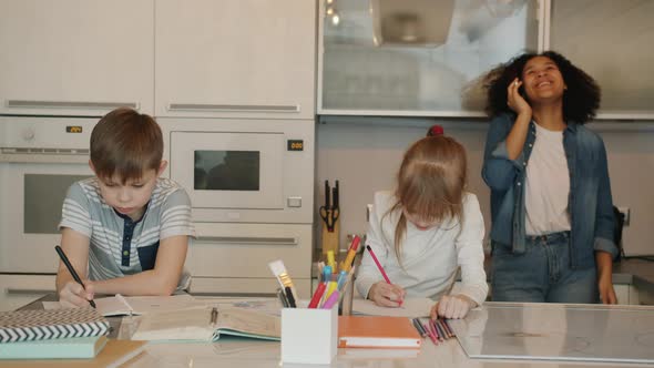 Joyful Babysitter Chatting on Mobile Phone and Laughing While Kids Doing Homework