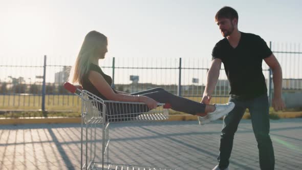 The Guy Rolls Attractive Young Woman in the Trolley on Parking Lot of the Supermarket at Sunset