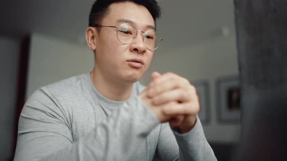 Asian serious man wearing eyeglasses talking by video call on laptop