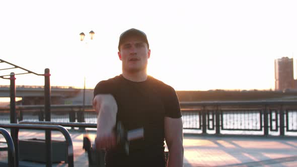 Young Sportive Man Training His Hands with Dumbbells Outdoors on Sunset  Looking in the Camera