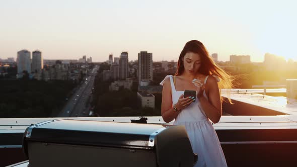 The girl is texting on the phone against the backdrop of a sunset on the roof of a skyscraper.