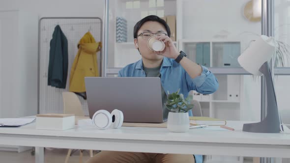 Asian Office Worker Working on Laptop and Drinking Coffee at Desk