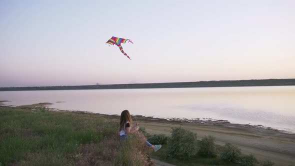 Happy Young Woman Playing with a Kite on a Glade with Lake Background at Sunset in Summer