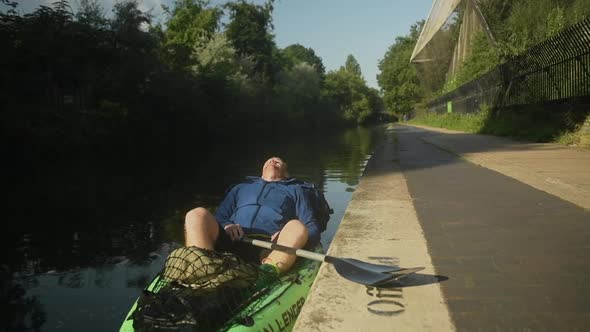 An adult man rests on the side of a river in his green kayak taking a moment to breathe in thoughtfu