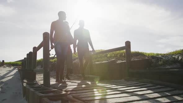 African american senior father and two teenage sons holding fishing rods walking on the beach