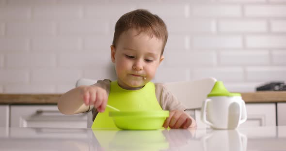 Zoom Shot of Small Cute Baby Boy Sitting at a Table and Eats Oatmeal with a Spoon