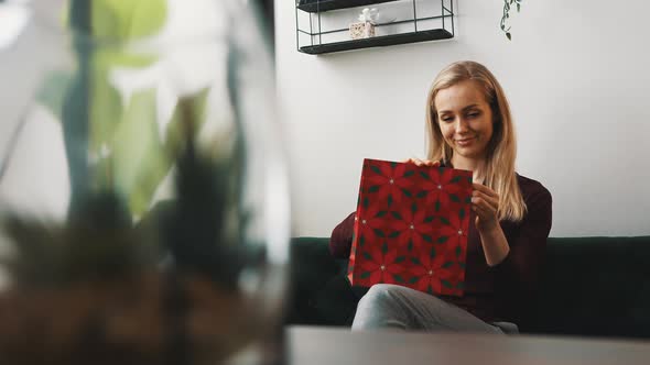 Beautiful Fairhaired Caucasian Woman Sitting in Her Living Room and Slowly Opening Red Gift Bag That