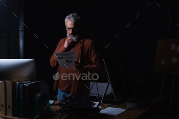 thoughtful businessman touching beard and looking at infographics at night in office