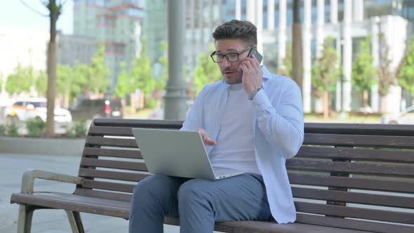 Man Talking on Phone and Using Laptop While Sitting Outdoor on Bench