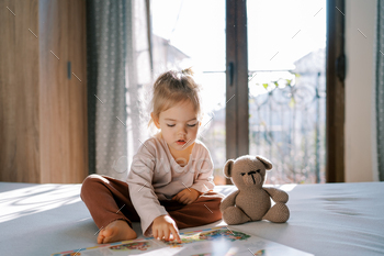 Little girl reads a book to a teddy bear sitting on the bed and running her finger across the page