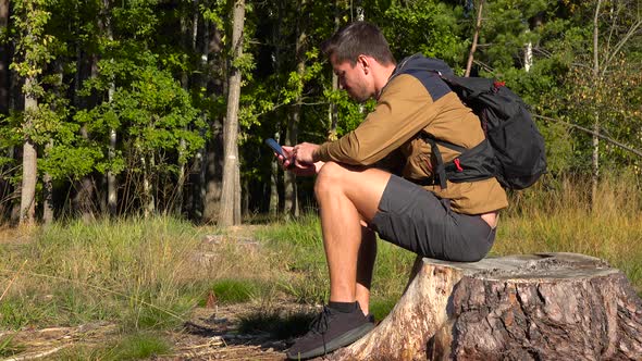 A Young Handsome Hiker Sits on a Tree Stump in a Forest and Works on a Smartphone