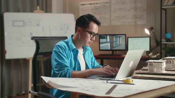 Asian Man Engineer Looking At Blueprint And Typing On A Laptop At The Office