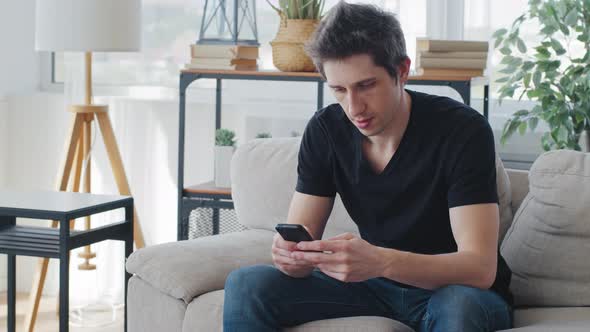 Serious Adult Caucasian Man Wears Black Tshirt Sitting at Home Sofa Living Room Looks at Mobile