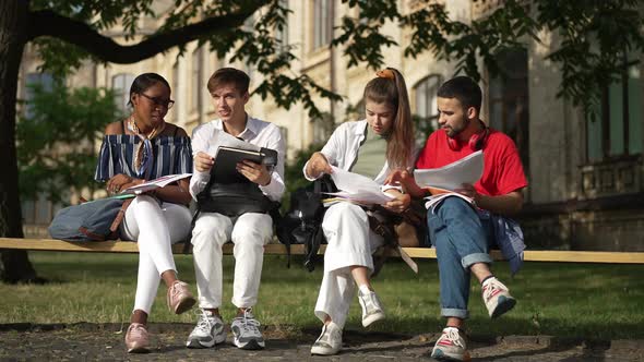 Wide Shot Front View Happy Young Students Sitting on Bench Talking and Studying Outdoors on Campus