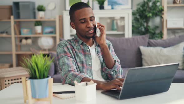 Cheerful African American Freelancer Is Talking on Mobile Phone and Using Laptop Working at Home