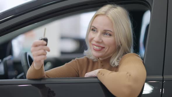 Happy Satisfied Blond Woman Bragging New Car Key Sitting on Driver's Seat in Showroom