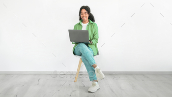 Smiling black woman sits with laptop computer against white wall