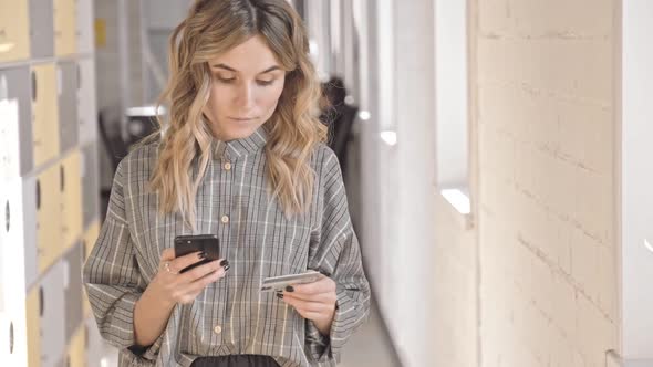 Serious Young Businesswoman Using Smartphone and Credit Card Walking in Office Room