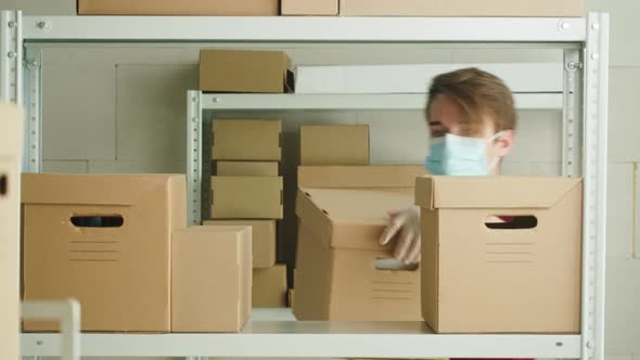 Man Wearing Medical Face Mask and Protective Gloves Putting Parcel on Shelf Employee of Warehouse