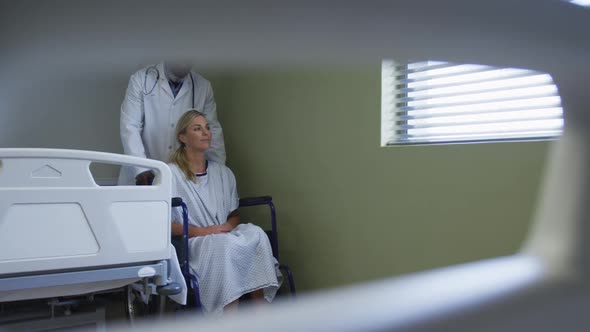 Diverse male doctor transporting female patient sitting in wheelchair in hospital room