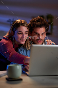 Young couple using a laptop together at home during nighttime searching for a travel destination.