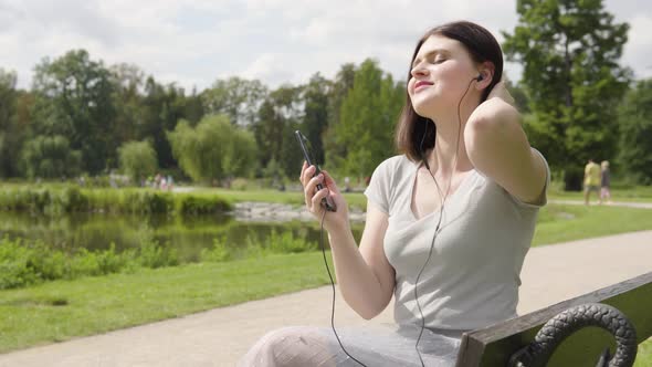 A Young Caucasian Woman Listens to Music with Earphones on a Smartphone As She Sits on a Bench