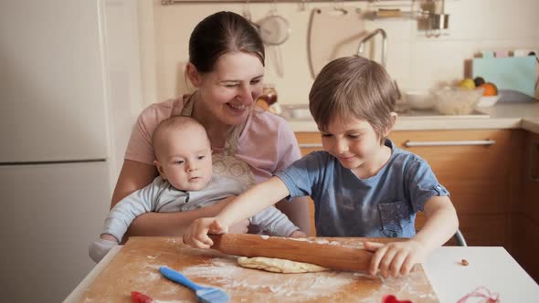 Young Mother with Little Baby Son Looking at Her Older Son Rolling Dough and Cooking