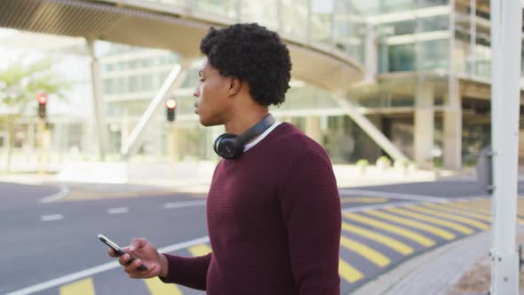 African american man in city, using smartphone, wearing headphones and backpack in street