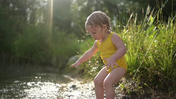 Little Funny Cute Blonde Girl Child Toddler in Yellow Wet Bodysuit Playing By the Lake Waterside