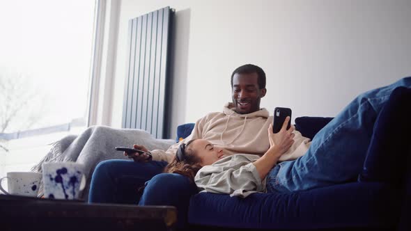 Relaxed Young Couple At Home Sitting On Sofa Watching TV And Checking Social Media On Mobile Phone