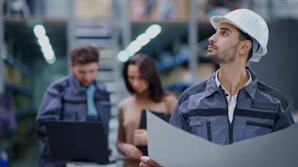 Handsome Male Warehouse Inspector Holding Paperwork Looking Up at Stock on Racks As Blurred Workers