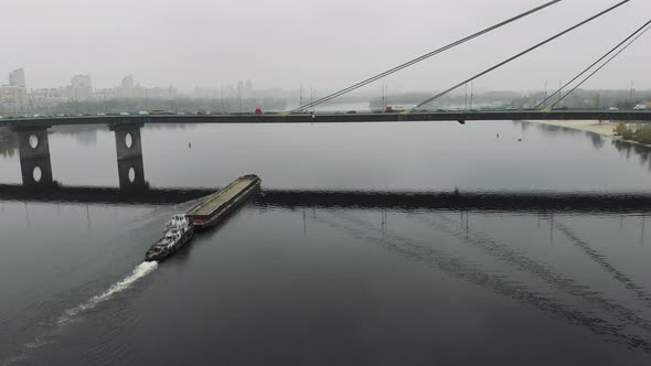 Barge laden with sand floating on empty calm river through loaded bridge in industrial downtown