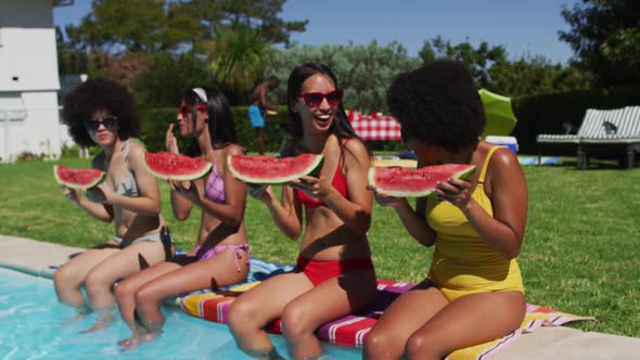 Diverse group of female friends eating watermelon sitting at the poolside talking