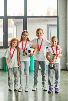 vertical shot of preadolescent cute children with golden medals posing with soccer ball and trophy