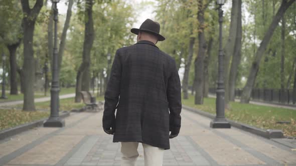 Back View of Elegant Man in Coat, Hat, and Gloves Walking Along Spring or Autumn Park. Camera