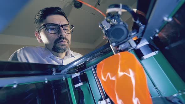 A Man Watches Printing Machine Creating an Orange Vase. Professional Industrial Designer Creates 3d