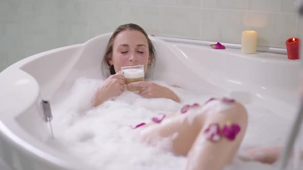 Slim Relaxed Caucasian Woman Lying in Bath Foam in Bathtub Smelling Tasty Cappuccino