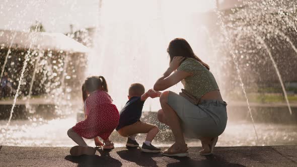 Little Girl Jumps Mother Holds Boy Touching Fountain Water
