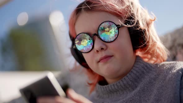 A Stylish Teenager with Pink Hair is Looking Through a Mobile Phone and Listening to Music with