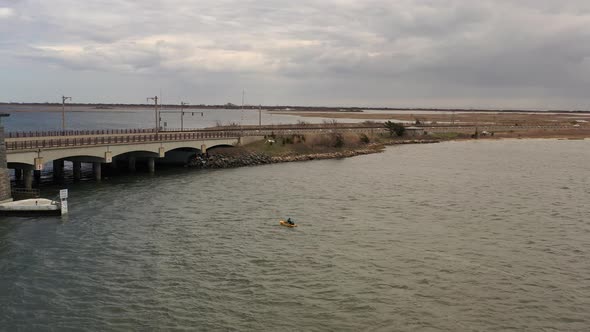 An aerial shot over a salt marsh on a cloudy day. There is a man in a yellow kayak paddling towards